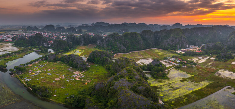 Aerial view of Tam Coc - Bich Dong featuring dramatic limestone karst formations, lush green rice paddies, winding waterways, and a vibrant sunset sky in Nam Hoa Lu, Ninh Binh, Vietnam.