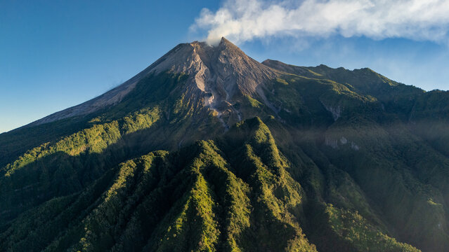 Aerial view of Mount Merapi with volcanic smoke rising from the crater, lush green slopes, and rugged terrain in Kecamatan Cangkringan, Daerah Istimewa Yogya, Indonesia.