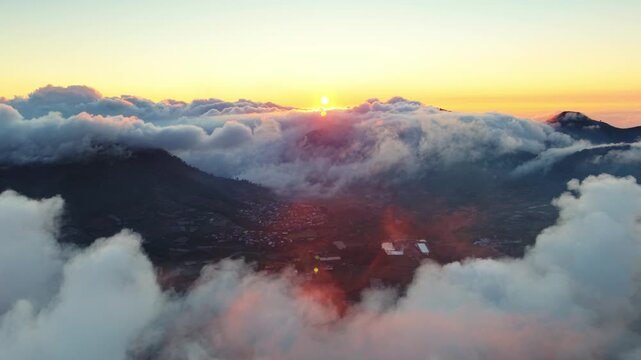 Aerial view of sunrise over Dieng Plateau with clouds blanketing the mountainous landscape and a small village nestled in the valley in Kecamatan Batur, Jawa Tengah, Indonesia.