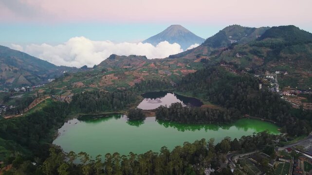 Aerial view of Dieng Plateau featuring the vibrant green Telaga Warna lake surrounded by lush volcanic hills and misty clouds in Kecamatan Batur, Jawa Tengah, Indonesia.