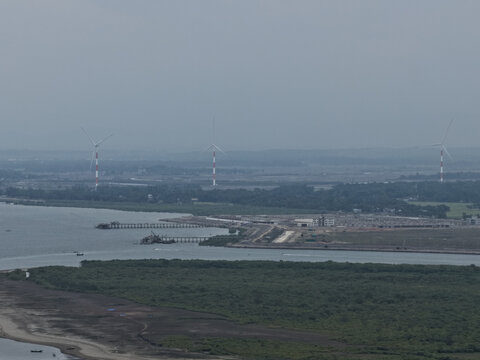 Cox's Bazar, Bangladesh - 09 April 2026: Aerial view of wind turbines standing in a coastal landscape with green mangroves, a river estuary, and industrial infrastructure.