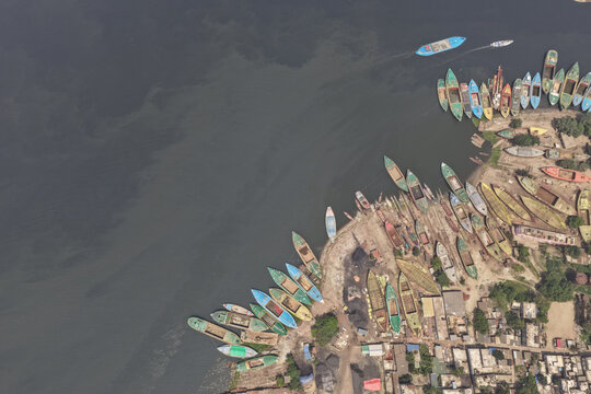 Dhaka, Bangladesh - 12 April 2026: Aerial view of numerous wooden cargo boats moored along the riverbank in the Buriganga River area, with industrial activity and urban structures visible nearby.
