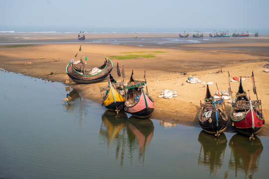 View of traditional wooden fishing boats moored on the sandy shore with a fisherman casting a net in the calm water in Cox's Bazar, Chittagong Division, Bangladesh.