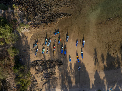 Aerial view of long-tail boats moored on the sandy shore of Secret Beach at low tide with rocky formations and palm tree shadows in Ko Lanta, Thailand.