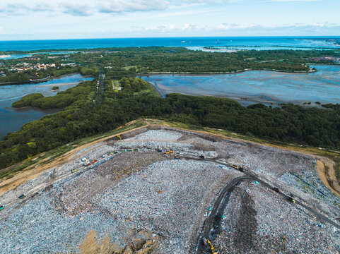 View of a large landfill site with heavy machinery operating amidst waste, surrounded by lush mangrove forests and the coastal landscape in Denpasar, Bali, Indonesia.