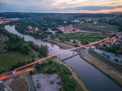 Aerial view of the illuminated Troja Footbridge spanning the Vltava River at sunset, with the historic Troja Palace and its manicured gardens visible in Prague, Czechia.