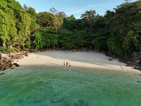 Aerial view of Phi Phi Islands beach with white sand, clear turquoise water, lush green trees, and three people standing on the shore in Mueang Krabi District, Krabi, Thailand.