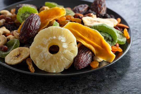Dried tropical fruits, nuts, and raisins in black plate.