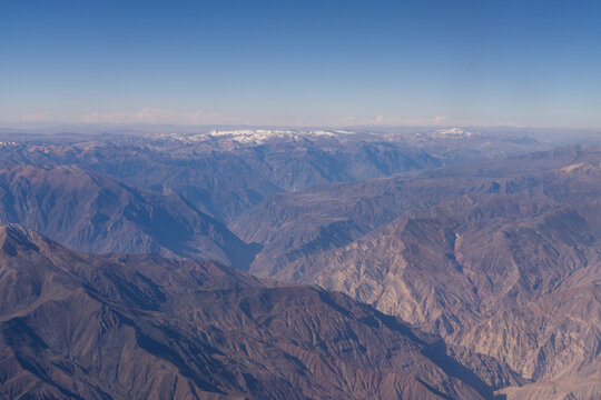 Aerial view of Colca Canyon featuring deep rugged valleys, steep mountain slopes, and distant snow-capped peaks under a clear blue sky in Arequipa, Peru.