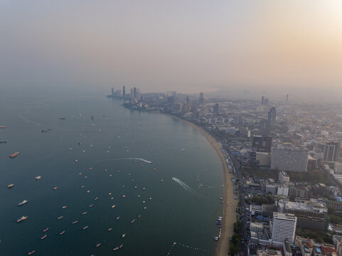 Aerial view of the Pattaya City coastline featuring high-rise buildings, a sandy beach, and numerous boats anchored in the bay during a hazy sunset in Pattaya City, Chon Buri, Thailand.