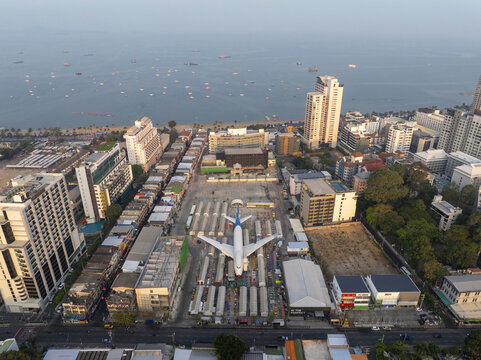 Aerial view of the airplane night market featuring a large aircraft fuselage surrounded by rows of market stalls and urban buildings near the coast in Pattaya City, Chon Buri, Thailand.