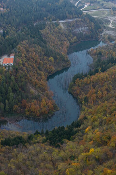 Aerial view of Lago di Poggio Baldi surrounded by autumn forests with vibrant foliage, featuring dead trees in the water and a building with a red roof in Santa Sofia, Emilia-Romagna, Italy.