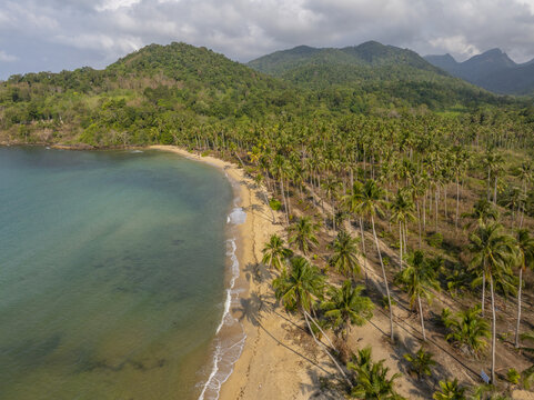 Aerial view of Wai Chaek Beach with a sandy coastline, lush palm trees, turquoise waters, and dense tropical mountains in the background in Koh Chang Tai, Trat, Thailand.