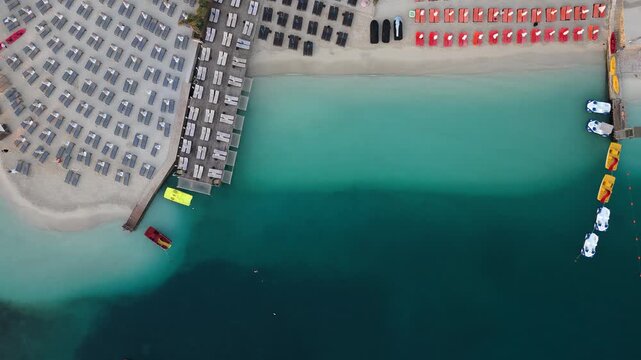 Aerial view of a beach with rows of sun loungers and umbrellas along the turquoise sea with pedal boats moored at the pier in Ksamil, Vlore County, Albania.