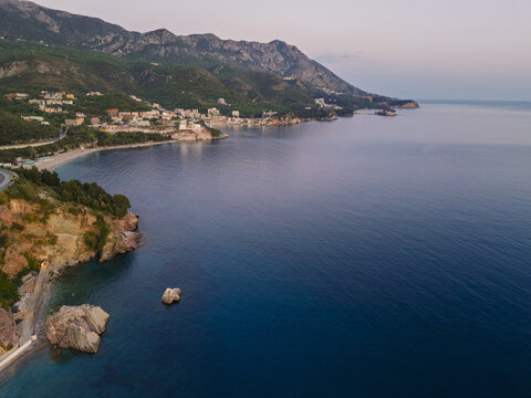 Aerial view of the coastal village of Rafailovici nestled along the Adriatic Sea with rugged mountains, rocky cliffs, and calm blue waters in Rafailovici, Opstina Budva, Montenegro.