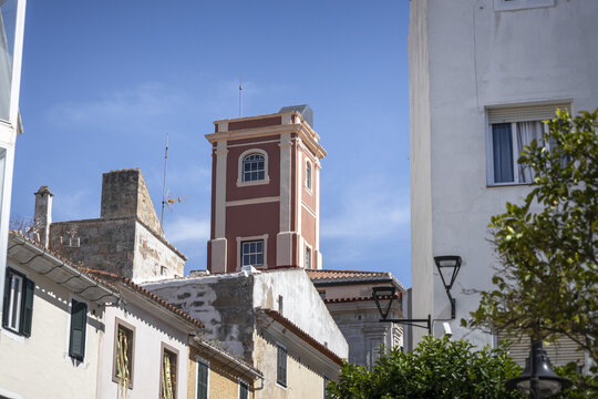 View of a distinctive red tower with arched windows rising above traditional Mediterranean buildings under a clear blue sky in Mao, Balearic Islands, Spain.