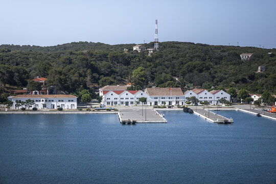 View of the historic naval base buildings and pier at the Port of Mahon, with a forested hillside and communication tower in the background in Mao, Balearic Islands, Spain.