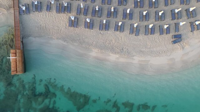 Aerial view of rows of sun loungers on the sandy beach with a wooden pier extending into the turquoise sea in Ksamil, Vlore County, Albania.