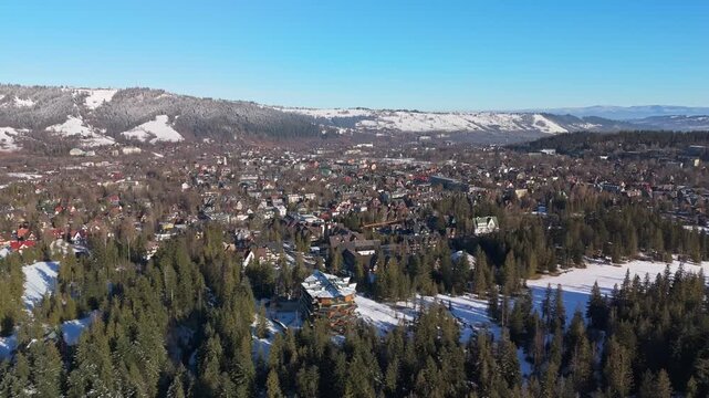 Aerial view of Zakopane in the Tatra Mountains, Poland, with snowy chalets, pine forest and ski resort buildings under a clear blue winter sky.