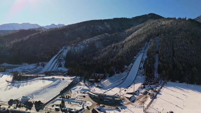 Aerial view of Zakopane ski jumping stadium in the Tatra Mountains, Poland, with snowy slopes, ski lifts, grandstands and pine forest under a clear winter sky.
