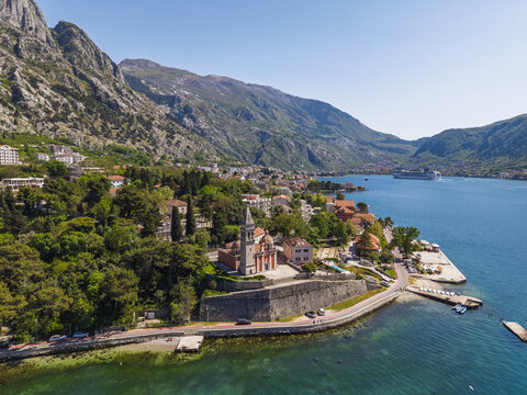 Aerial view of the Church of St. Matthew surrounded by lush greenery, stone walls, and the blue waters of the Bay of Kotor in Dobrota, Kotor Municipality, Montenegro.