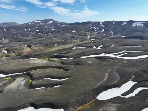 Aerial view of the volcanic landscape of Raudufossafjoll with dark lava fields, patches of snow, and mossy green terrain under a clear blue sky in Hella, Rangarthing ytra, Iceland.