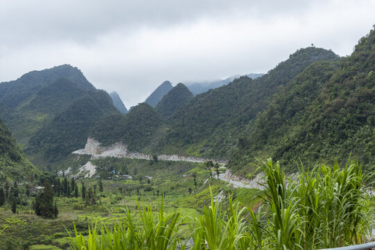 View of the winding Ha Giang Loop road cutting through lush green mountains under a cloudy sky in Dong Van, Tuyen Quang, Vietnam.