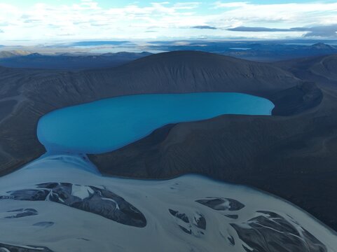 Aerial view of the vibrant blue Skyggnisvatn crater lake surrounded by dark volcanic terrain and glacial river channels in Hella, Rangarthing ytra, Iceland.