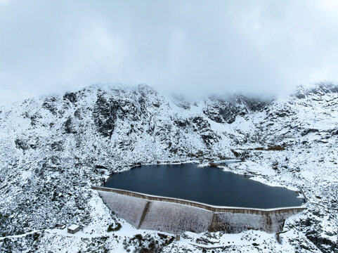 Aerial view of Barragem de Covao do Ferro surrounded by snow-covered mountains and rocky terrain in Unhais da Serra, Castelo Branco, Portugal.