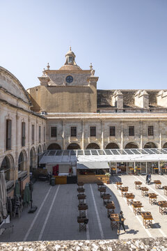 Mao, Balearic Islands, Spain - 25 July 2026: View of the historic Claustre del Carme courtyard featuring arched stone walkways, a central square with tables, and a church dome under a clear blue sky.