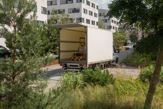 White delivery truck with tailgate open in a parking lot with modern buildings and green vegetation. Seen from side and back.