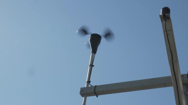 A cup anemometer spinning rapidly from the wind against a clear blue sky. Static handheld camera shot of a meteorological weather instrument in motion.