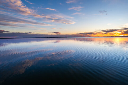 Aerial view of Lake Ellesmere Te Waihora at sunset, with calm water reflecting the colorful sky and soft clouds in the Canterbury Region, New Zealand.