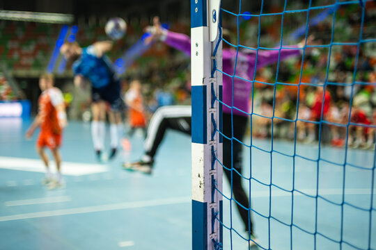 A view from behind a handball goalkeeper in a red jersey with arms outstretched, watching an opponent prepare for a shot during an indoor match.