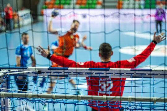Professional handball match action seen through the goal net, featuring a blurred goalkeeper and an attacking player jumping for a shot in a modern sports hall.