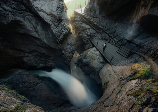 View of a person walking on a metal walkway through the narrow rocky gorge of Trummelbach Falls with cascading water and steep stone walls in Switzerland.