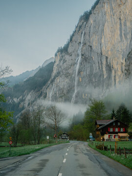View of the Staubbach Falls cascading down a massive cliff face in the Lauterbrunnen Valley, with a traditional wooden house and a road in the foreground in Switzerland.