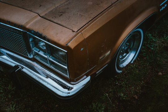 View of a weathered vintage brown car parked on grass, highlighting the chrome bumper, headlight, and classic wheel design in Port Angeles, Washington, United States.