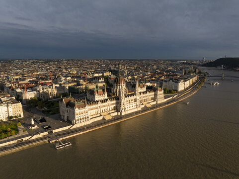 Aerial view of the Hungarian Parliament Building along the Danube River in Budapest, Hungary, featuring its iconic neo-Gothic architecture, red-tiled roof, and surrounding urban cityscape.