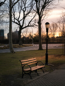 View of a wooden park bench situated on a grassy lawn next to a tall lamp post with bare trees and a sunset sky in the background, United States of America.
