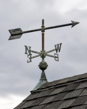 Old weathered metal directional vane wind indicator on a dark shingle roof against a cloudy sky
