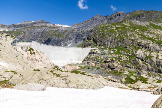 View of the VerticAlp Emosson dam set against rugged mountain peaks with rocky slopes, patches of snow, and lush green vegetation in Le Chatelard, Valais, Switzerland.
