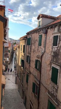 Narrow street in the historic old town of Sibenik, Croatia, with traditional stone buildings, green shutters and the Croatian flag waving in the sky