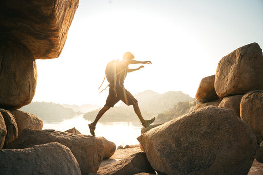 View of a hiker with a backpack jumping between large granite boulders during golden hour in the rocky landscape of Hampi, Karnataka, India.