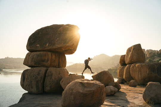 View of a person jumping between large granite boulders near the river during sunset in Hampi, Karnataka, India.