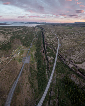 Aerial view of Thingvellir National Park featuring the Almannagja tectonic rift, winding roads, and mossy volcanic landscape under a soft sunset sky in Selfoss, Blaskogabyggd, Iceland.