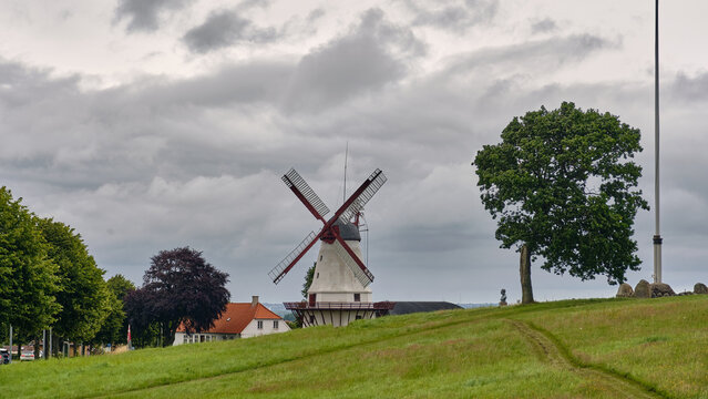 Sonderborg, Denmark - 06 July 2026: View of Dybbol Mill standing on a grassy hill under a cloudy sky with a large tree nearby.