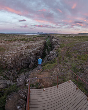 Aerial view of Thingvellir National Park with a person on a wooden platform overlooking the rugged volcanic gorge under a pink sunset sky in Selfoss, Blaskogabyggd, Iceland.