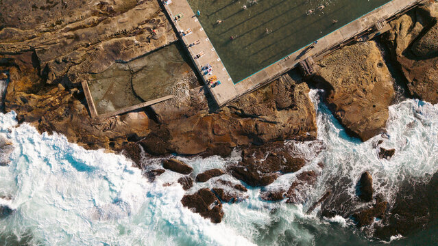 Aerial view of Dee Why Rock Pool with people swimming in the clear water and ocean waves crashing against the rocky coastline in Dee Why, New South Wales, Australia.