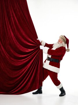 Santa claus pulling back a large red velvet curtain to reveal something, isolated on white background